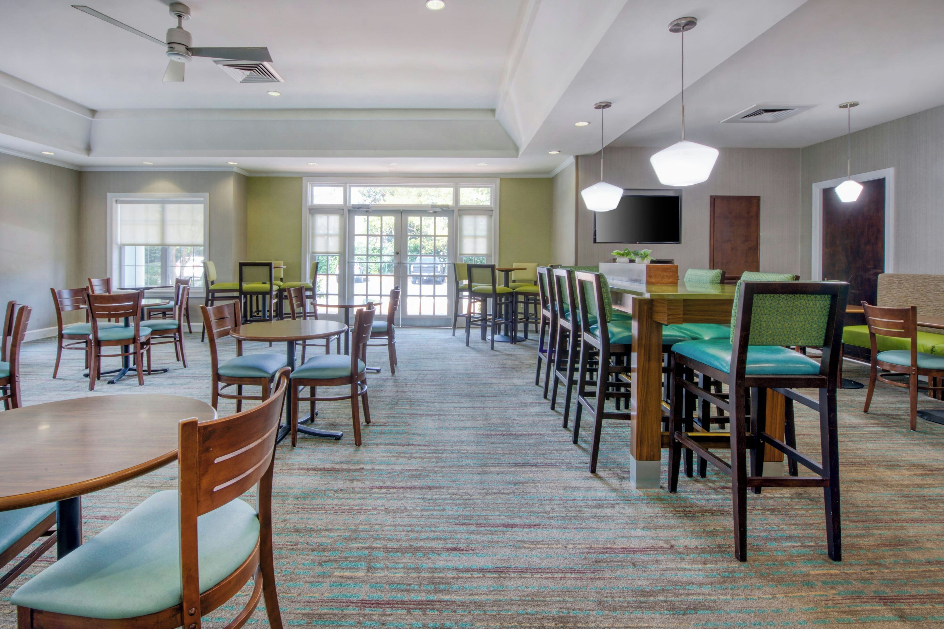 Een restaurantgedeelte van een hotel met meerdere tafels en stoelen, een flatscreen televisie aan de muur en een groot raam dat voor natuurlijk licht zorgt.