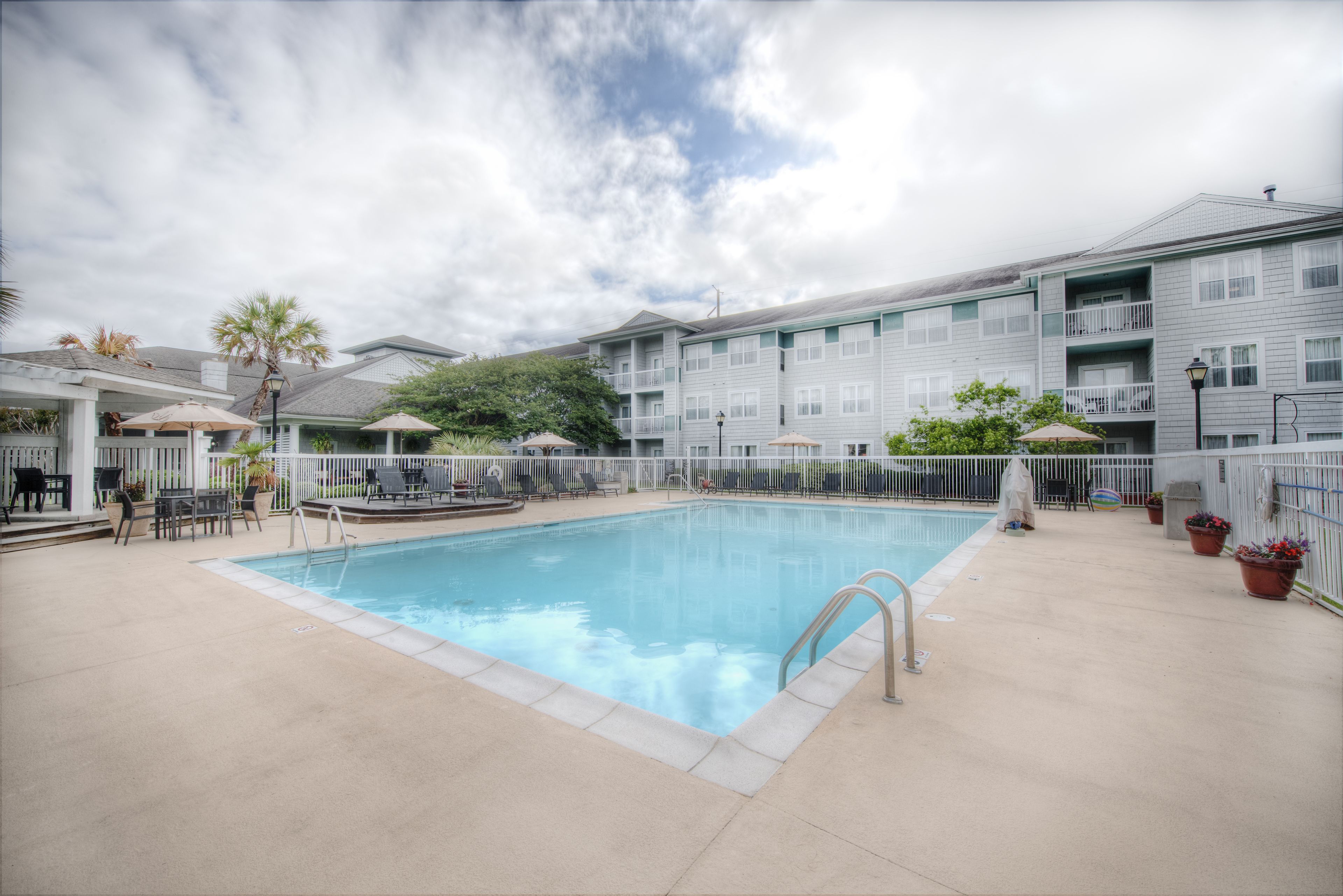 Una piscina rectangular con escalera metálica, rodeada por una valla blanca, contigua a un edificio de varios pisos con balcones.