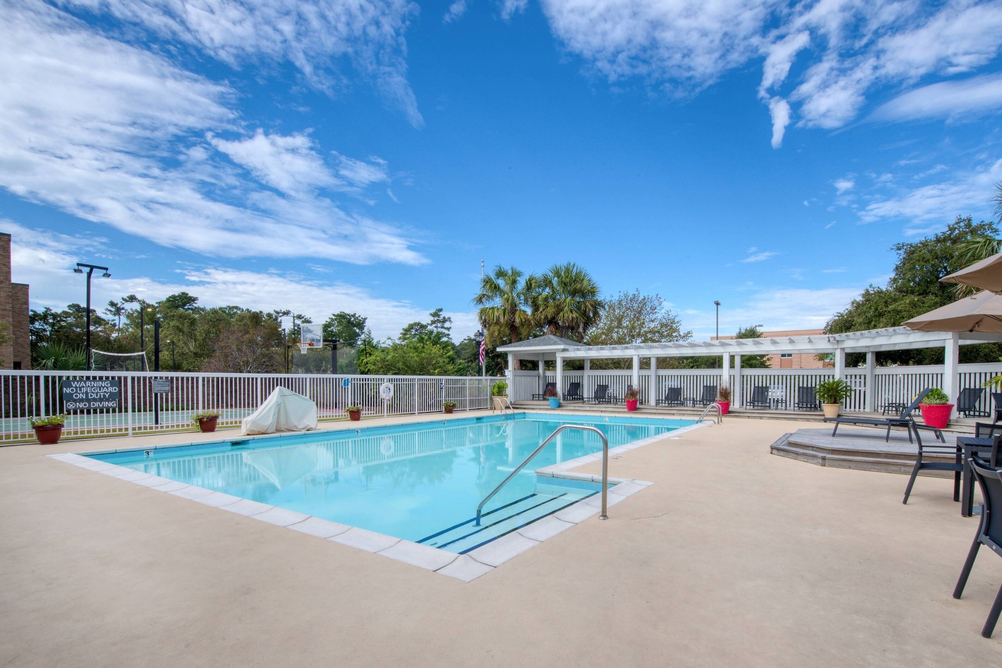 A swimming pool with a white fence, lounge chairs, and a covered seating area.
