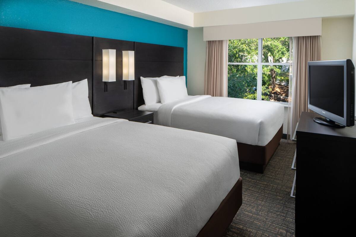 A hotel room with two beds, a TV, and a window with a view of trees.