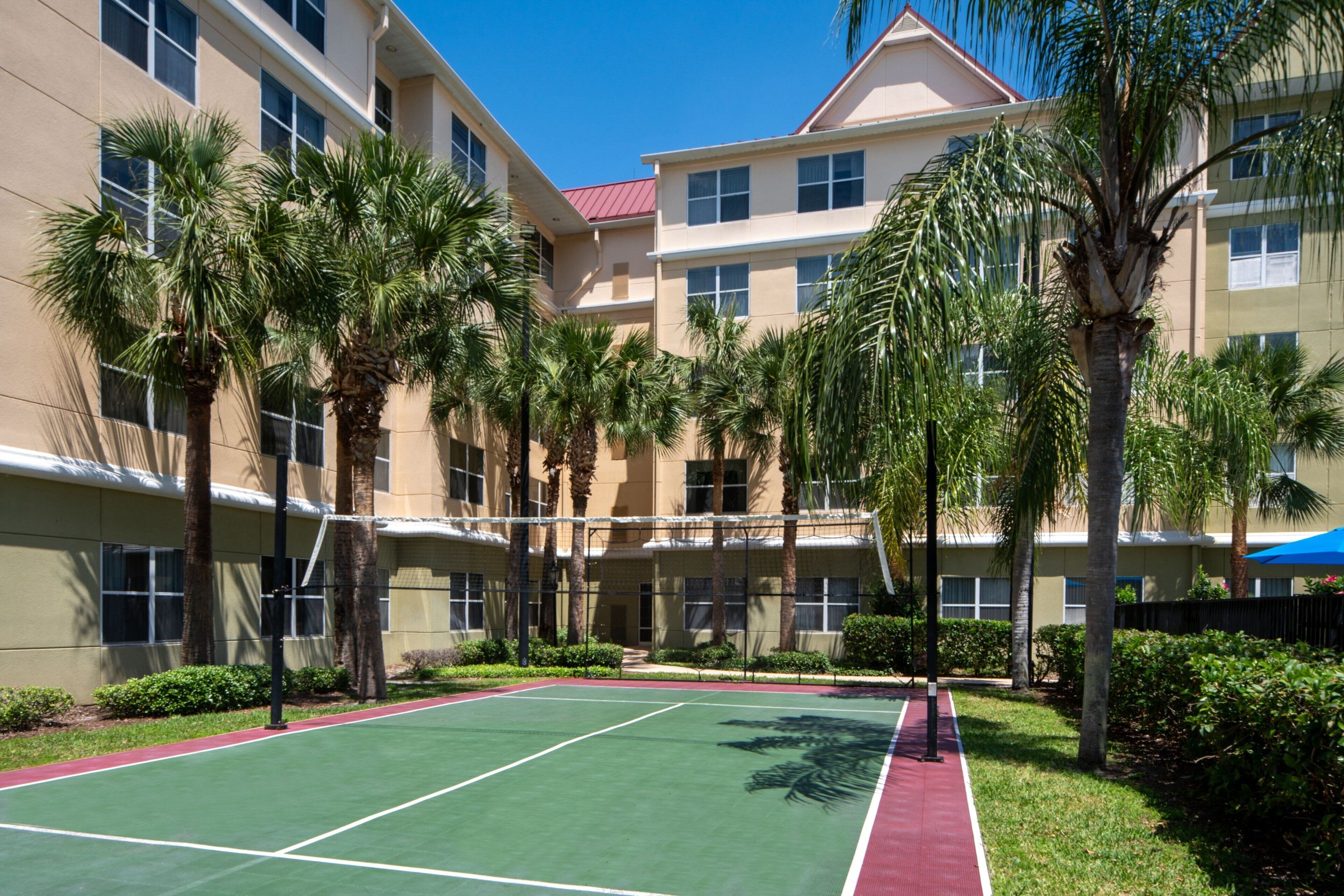 Una cancha de tenis rodeada de palmeras y un edificio de hotel de varios pisos con grandes ventanales.