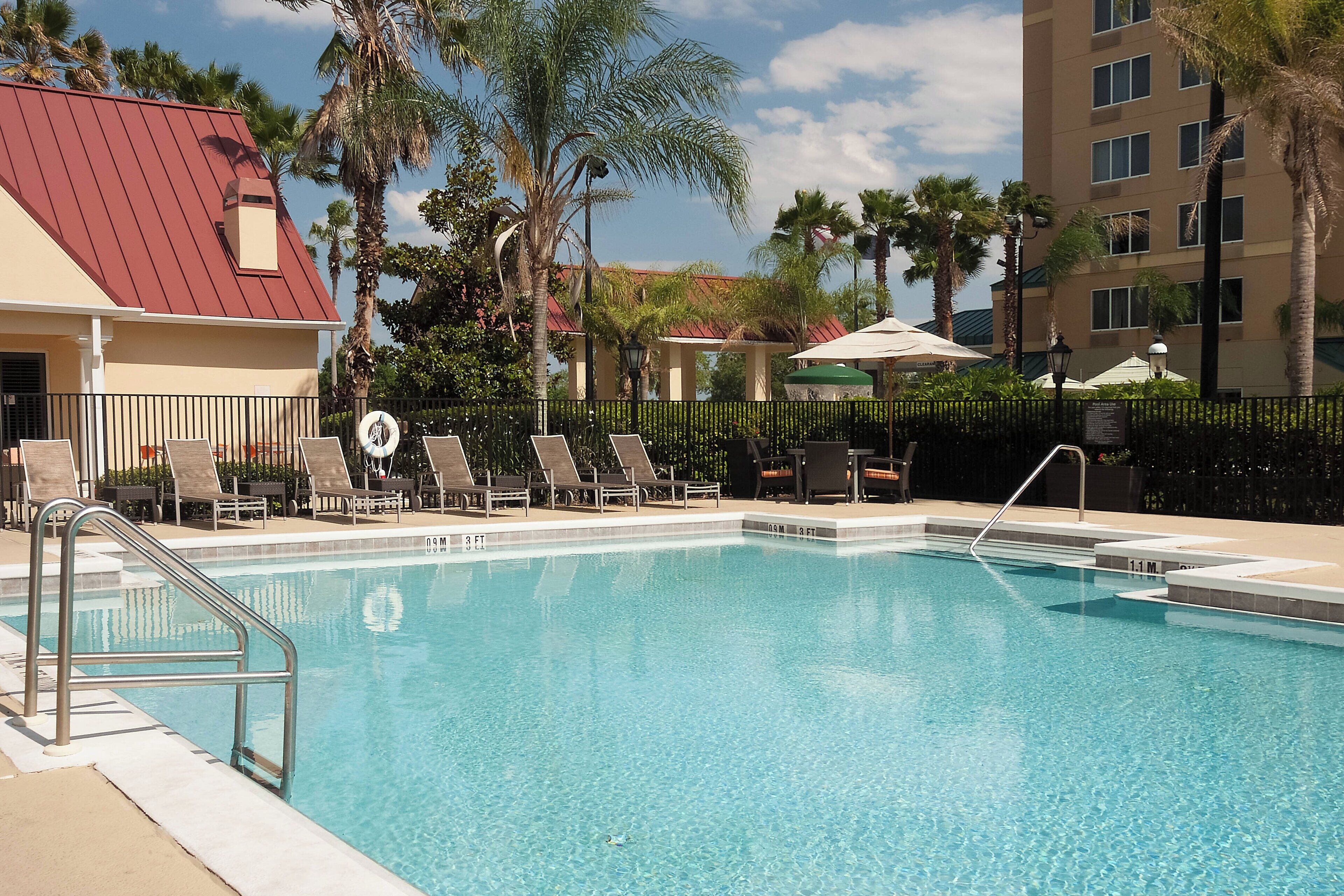 A swimming pool with lounge chairs and umbrellas, surrounded by palm trees and a building with a red roof.