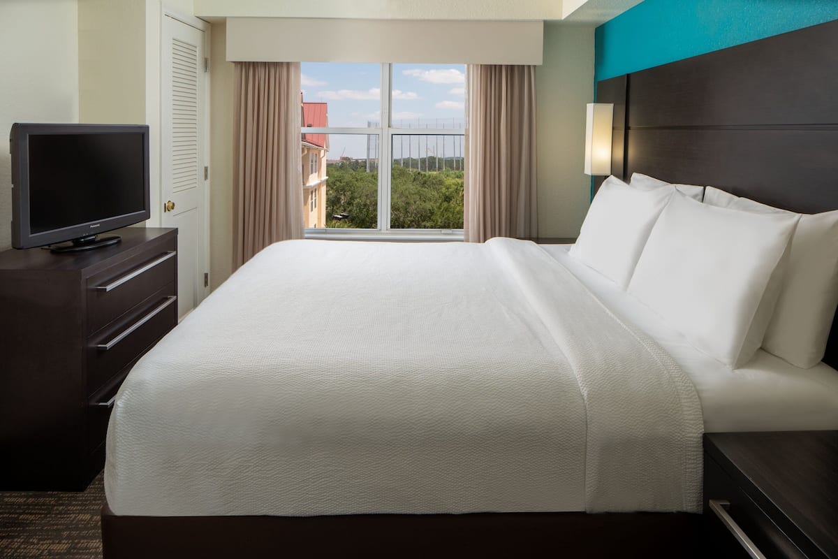 A hotel room with a large bed, a flat-screen TV on a dresser, and a view of buildings and trees through a window.