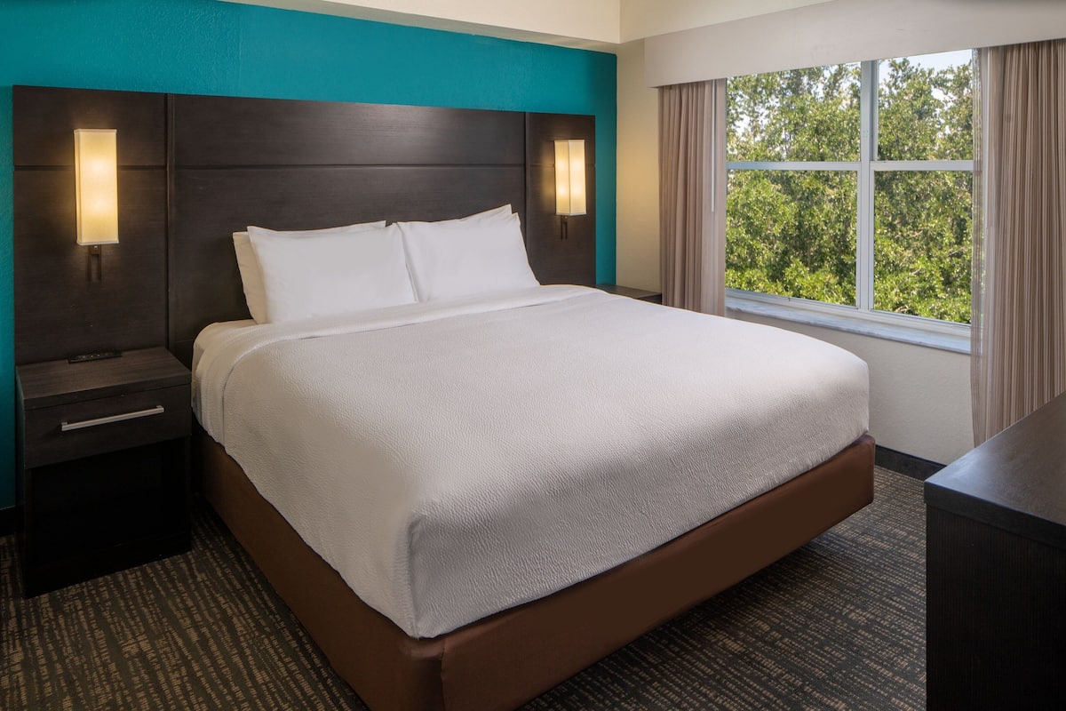 A hotel room with a large bed, two bedside tables, a nightstand, a window with curtains, and a view of trees.