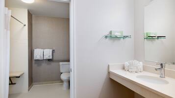 Baño con inodoro, lavamanos y espejo. Hay toallas y un estante de vidrio con artículos de aseo.