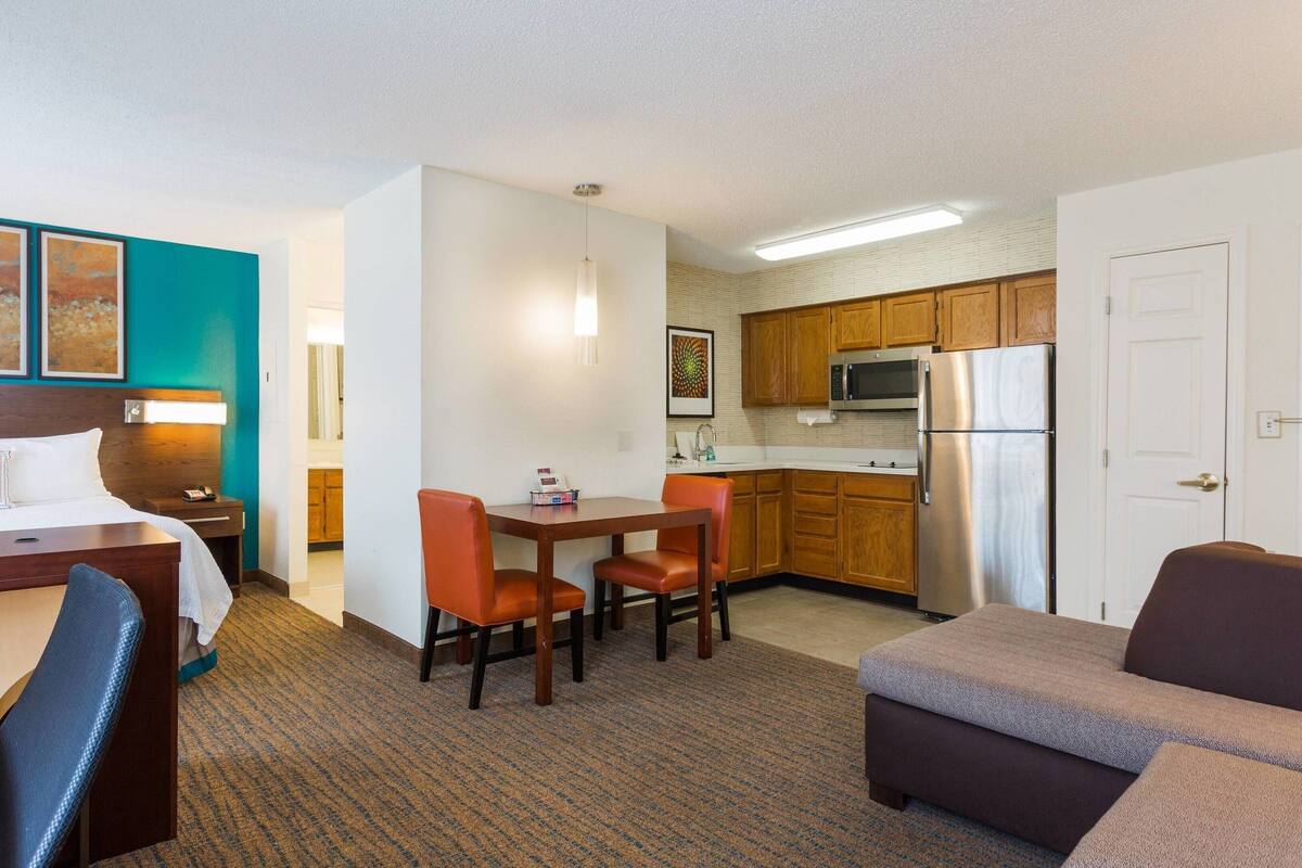 A hotel room with a bed, a desk, a dining table with chairs, a kitchenette with a refrigerator, and a TV.