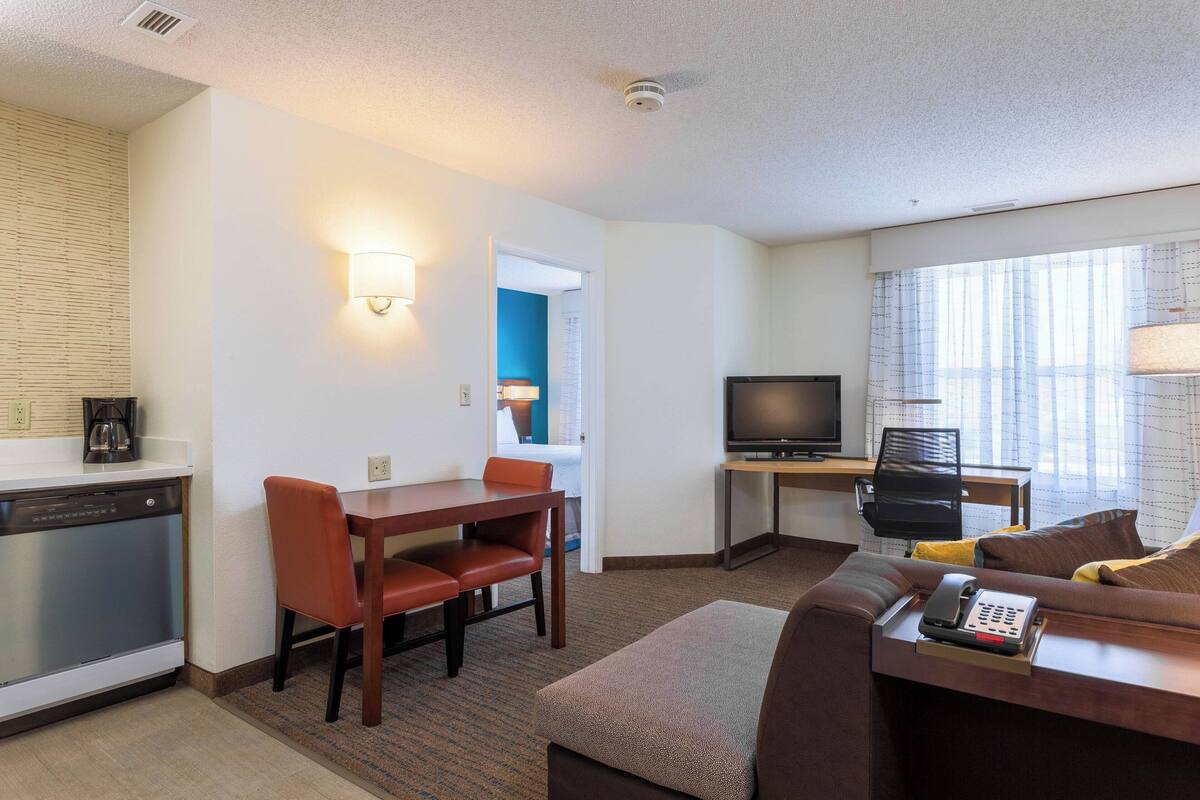 A hotel room with a kitchenette, a dining area with a table and chairs, a sofa with a phone, a TV, and a window with curtains.
