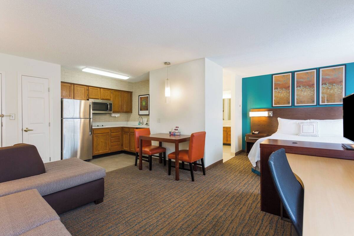 A hotel room with a kitchenette, a dining area with a table and chairs, a bed with a headboard, and artwork on the wall.