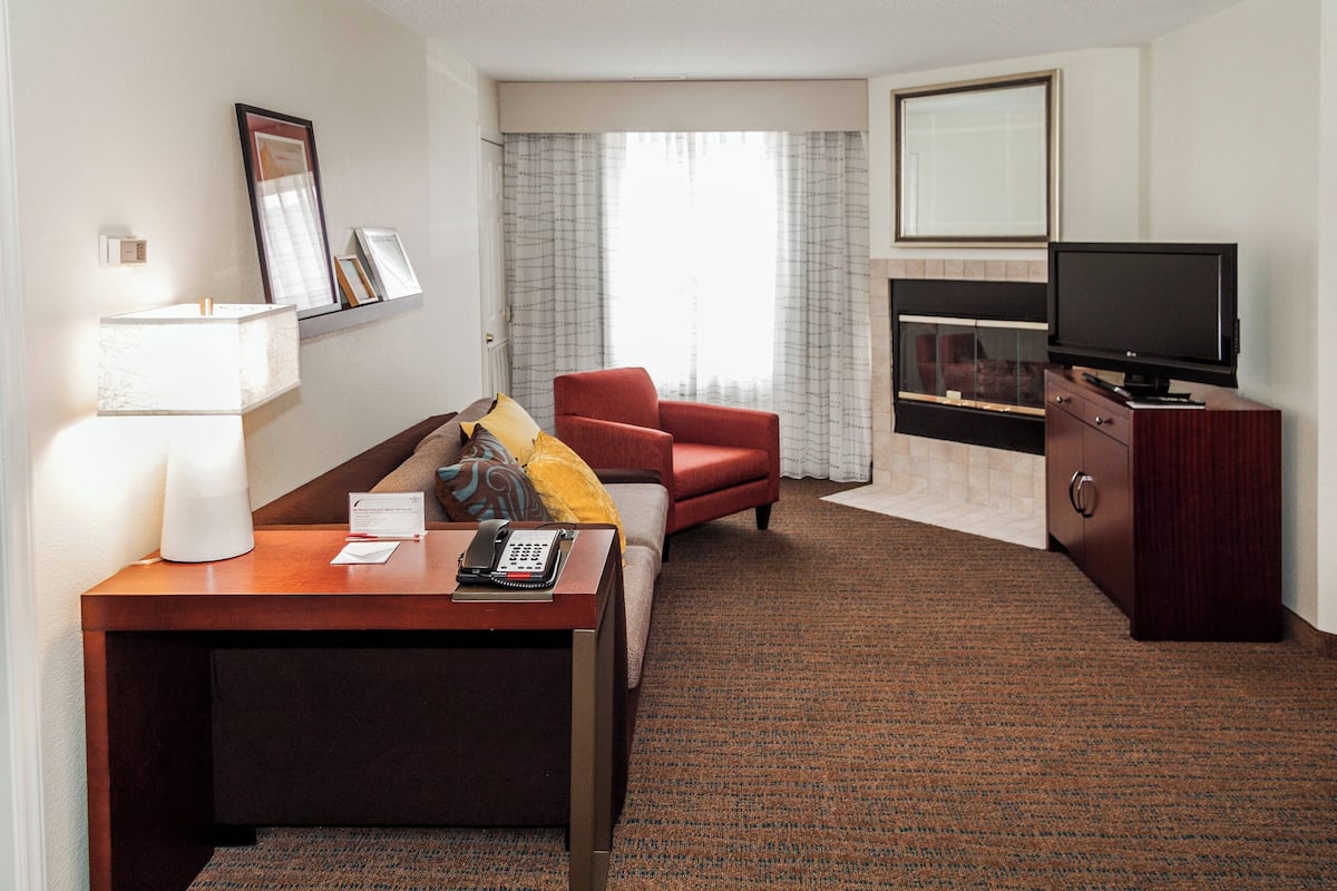 A hotel room with a wooden desk, a red armchair, a television, and a fireplace.