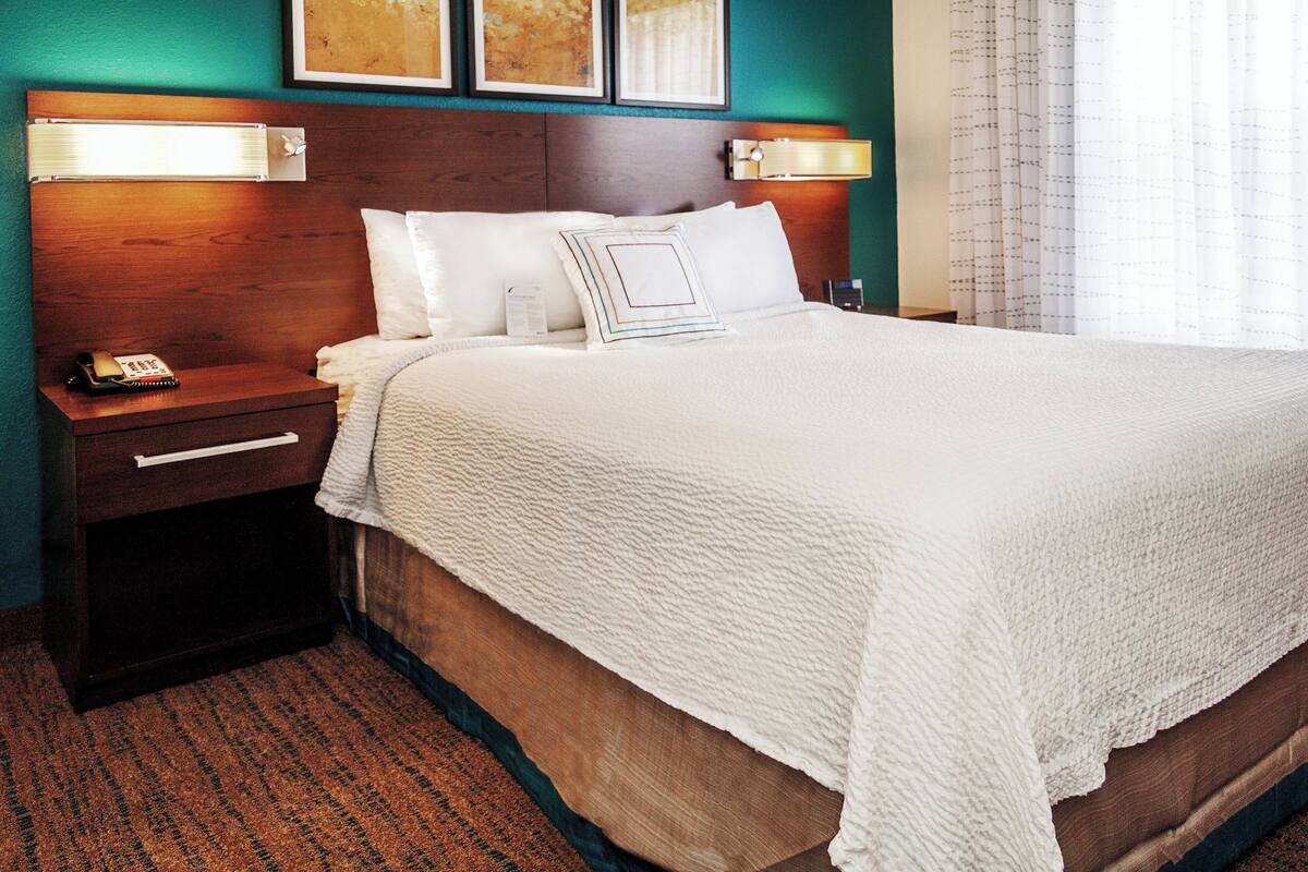 A neatly made bed with a white comforter and pillows, a wooden nightstand with a telephone, and a wall-mounted lamp.