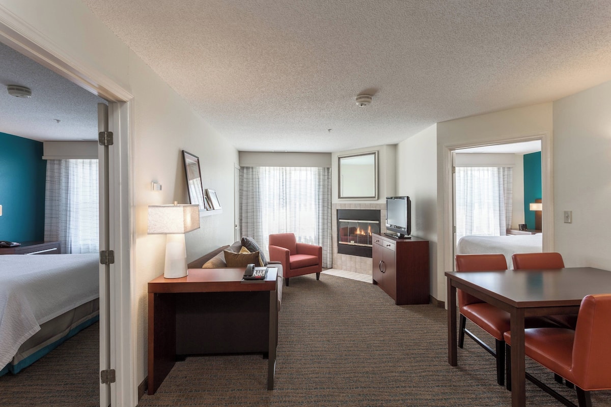 A hotel room with a bed, desk, chairs, a TV, and a fireplace.