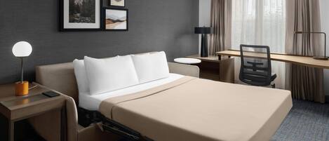 A hotel room with a bed, desk, chair, lamp, and framed pictures on the wall.