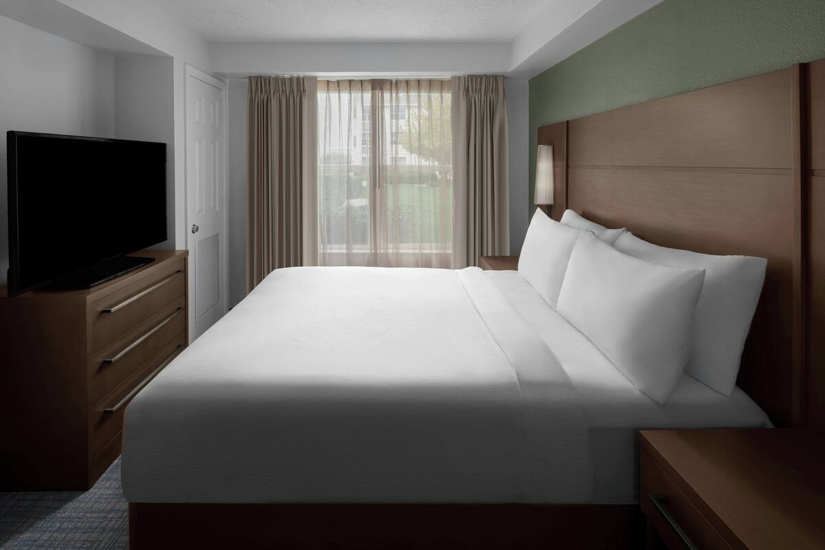 Une chambre d’hôtel avec un grand lit, un téléviseur posé sur une commode en bois et une fenêtre avec des rideaux.