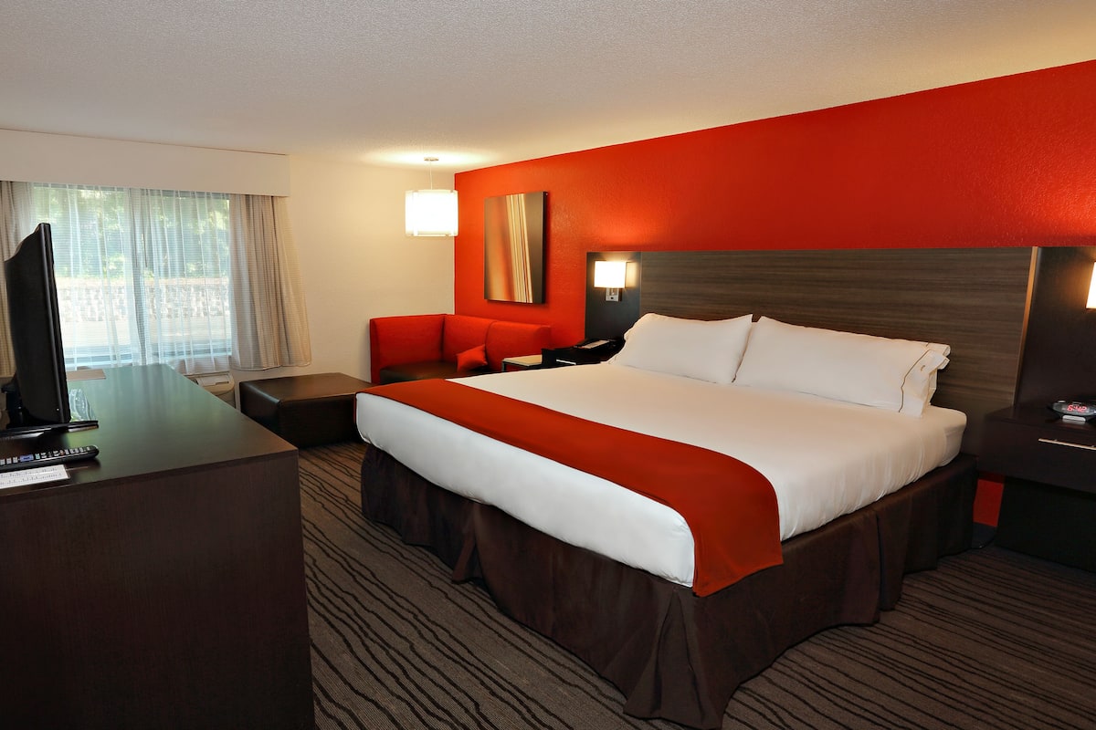 A hotel room with a large bed, a desk with a TV, a red armchair, and a nightstand with a lamp.