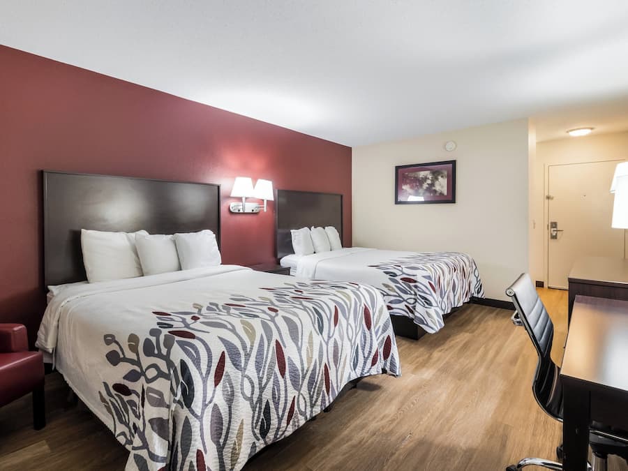 Habitación de hotel con dos camas, un escritorio, una silla, un sofá rojo y una lámpara de pared.