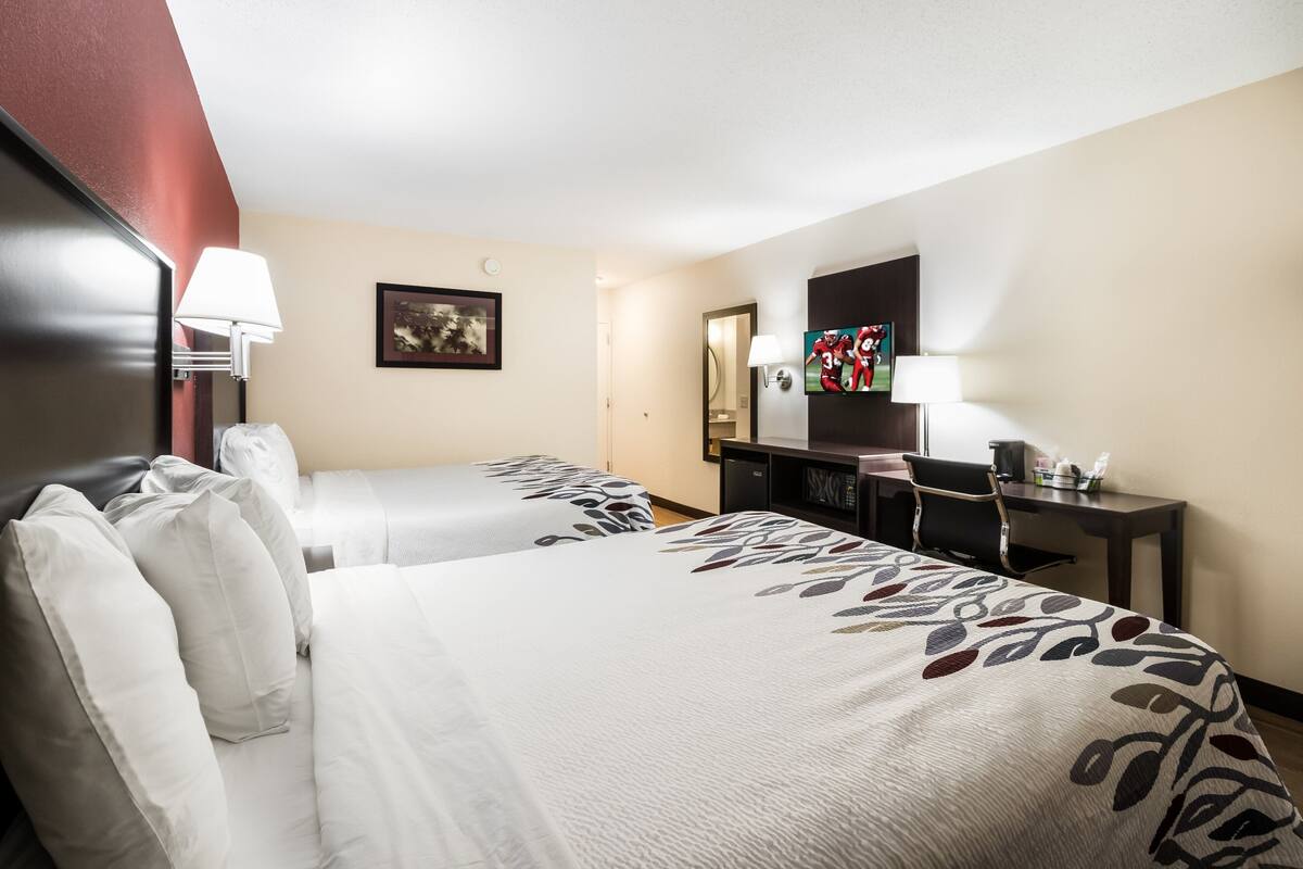 A hotel room with two beds, a desk, a chair, a lamp, and a television.