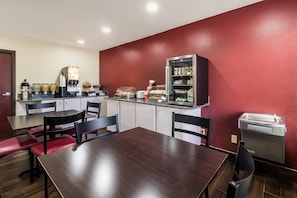 A restaurant with a red wall, a wooden table, and chairs. There is a coffee machine and a refrigerator with food items. - Red Roof Inn Murfreesboro (Murfreesboro)