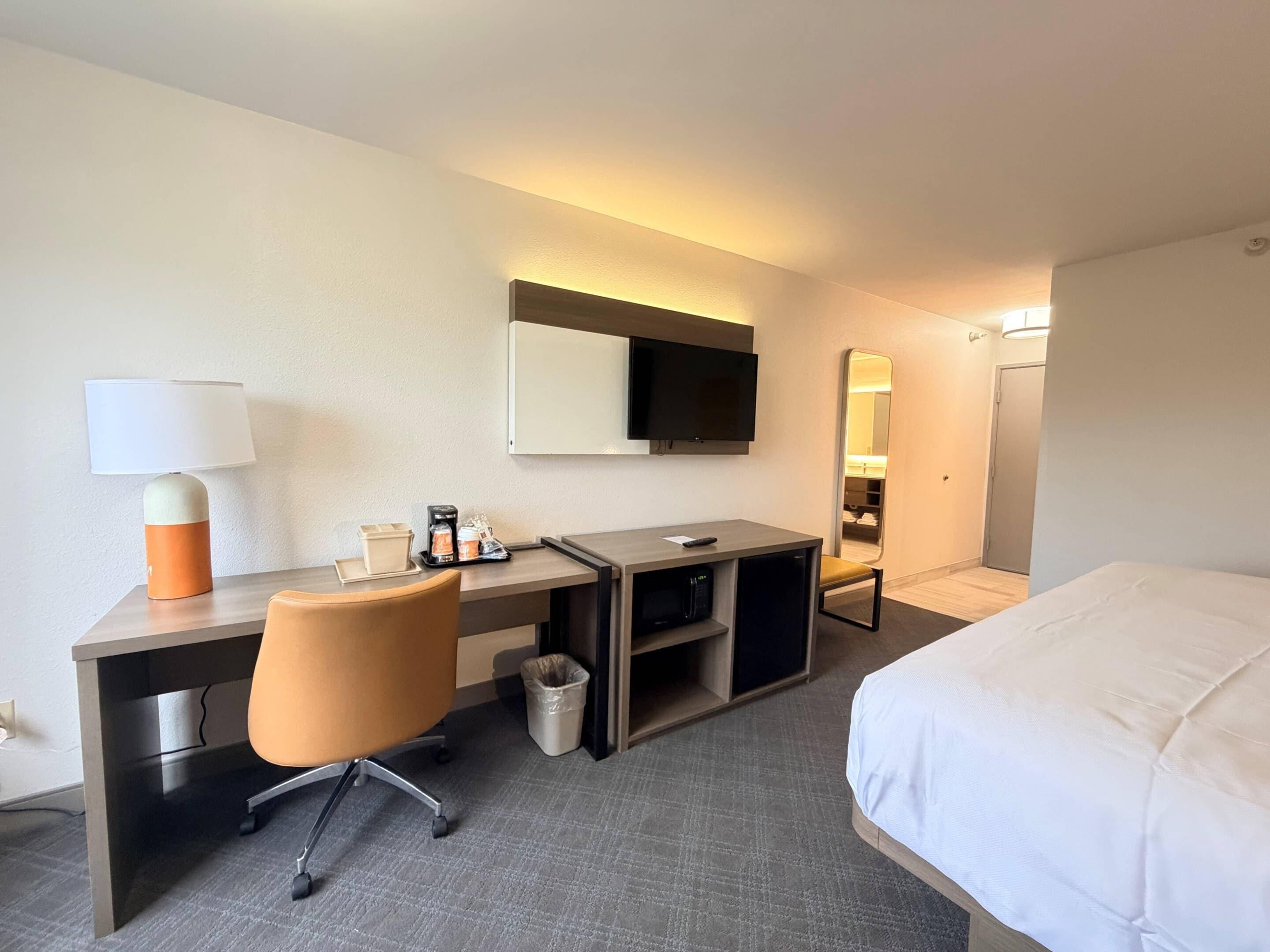 Ett hotellrum med en säng, ett skrivbord, en stol, en tv och en spegel.