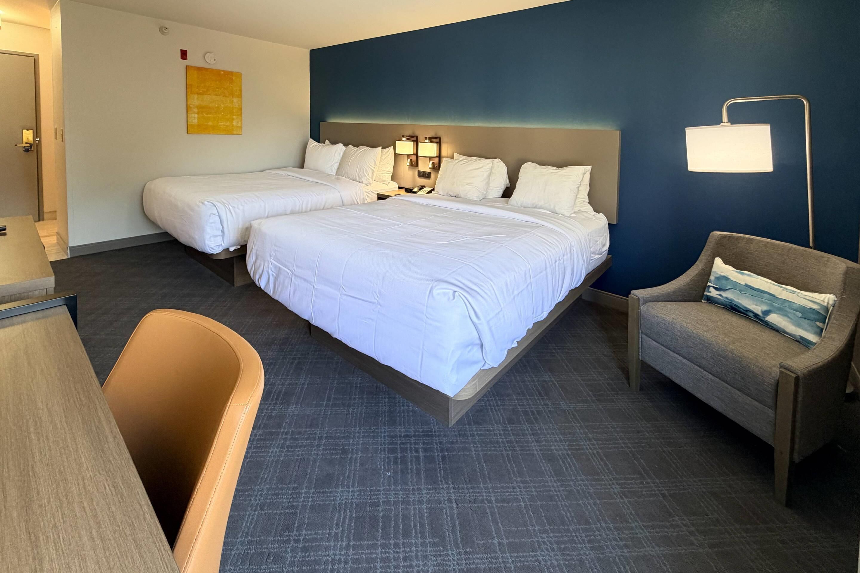 A hotel room with two beds, a desk, a chair, and a lamp.