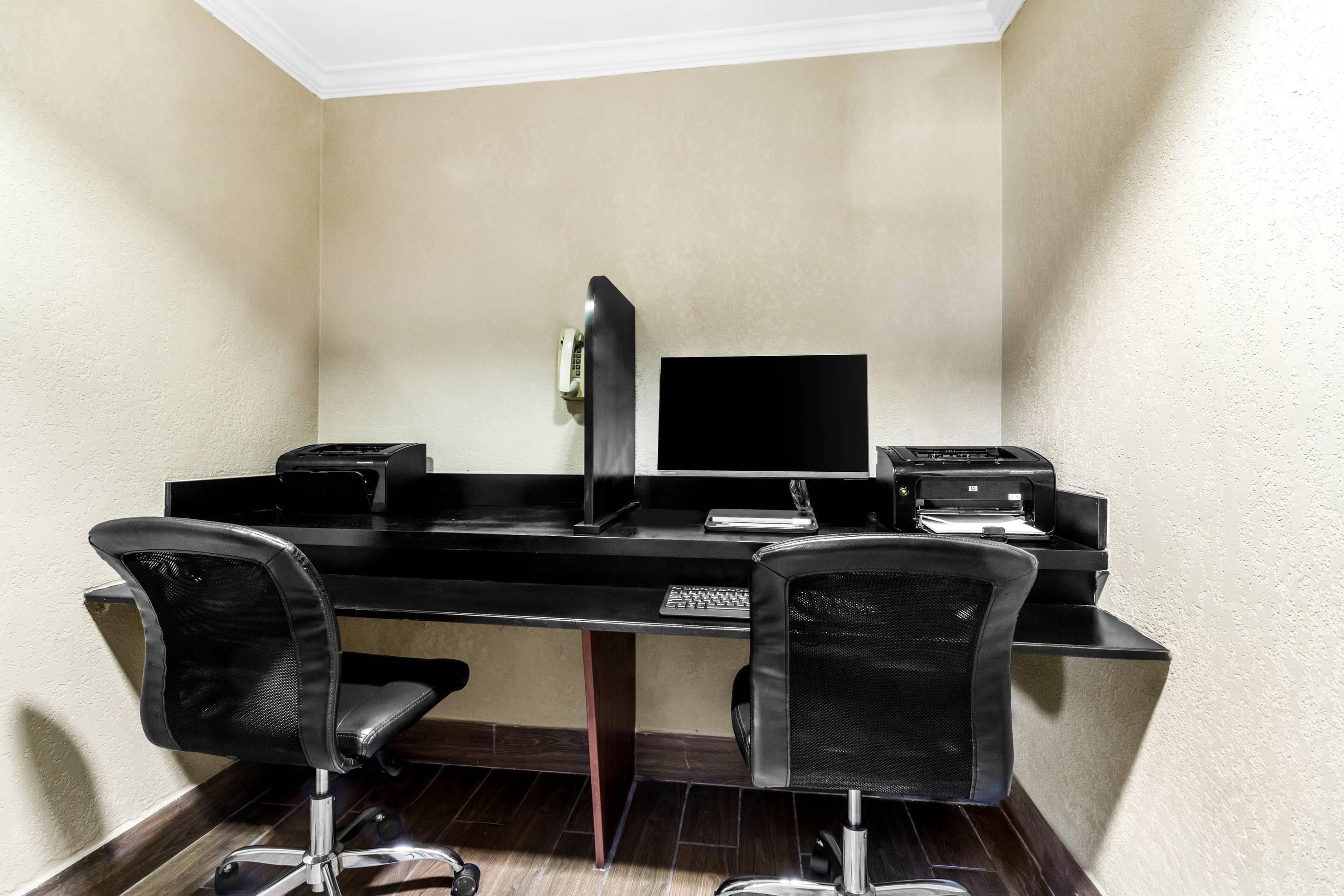 A corner office with a desk, two chairs, a computer monitor, a printer, and a telephone.