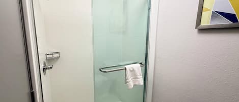A glass-enclosed shower stall with a towel rack, a glass door, and a white toilet visible in the adjacent bathroom.