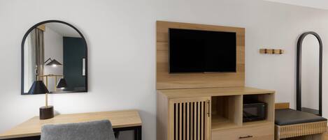 A hotel room with a wooden desk, a grey chair, a flat-screen TV mounted on the wall, a wooden wardrobe with a mirror, and a small bedside table with a lamp.