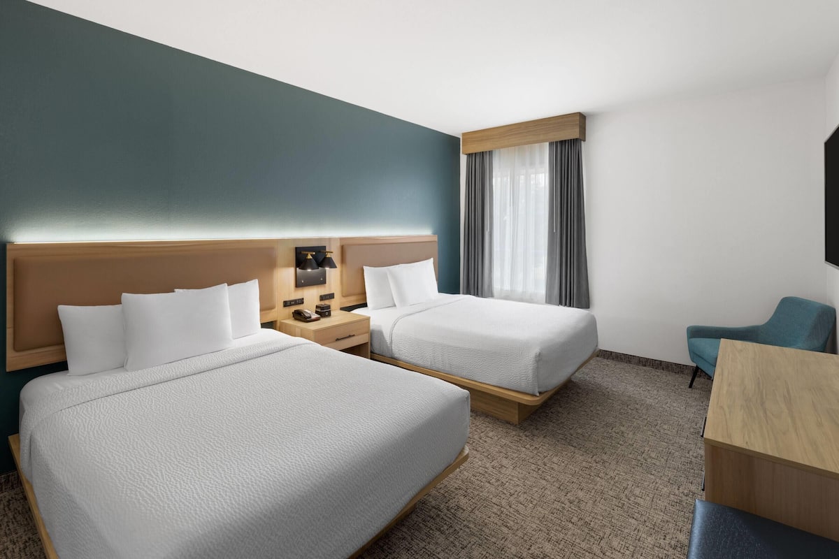 Une chambre d’hôtel avec deux lits, un bureau, une chaise, une table de chevet avec une lampe et une fenêtre avec des rideaux.
