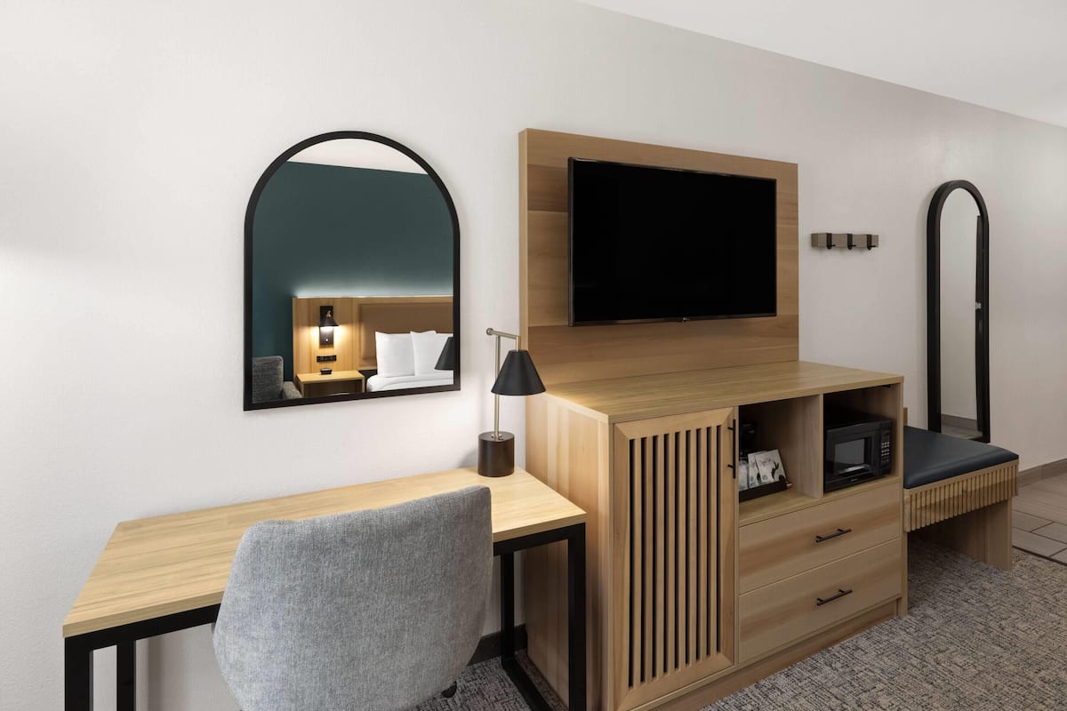 Une chambre d’hôtel moderne avec un bureau en bois, une chaise grise, un téléviseur à écran plat fixé au mur, une petite table de chevet avec une lampe et un four à micro-ondes.