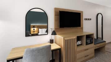 A modern hotel room with a wooden desk, a grey chair, a flat-screen TV mounted on the wall, a small bedside table with a lamp, and a microwave.
