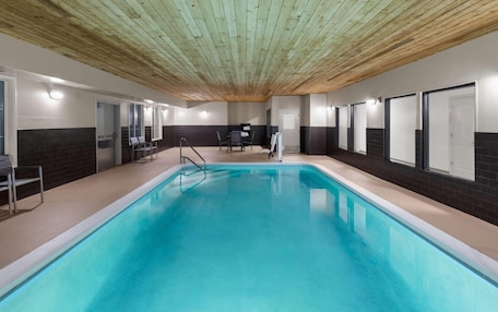 An indoor swimming pool with a wooden ceiling, tiled walls, and a seating area with chairs and a table.