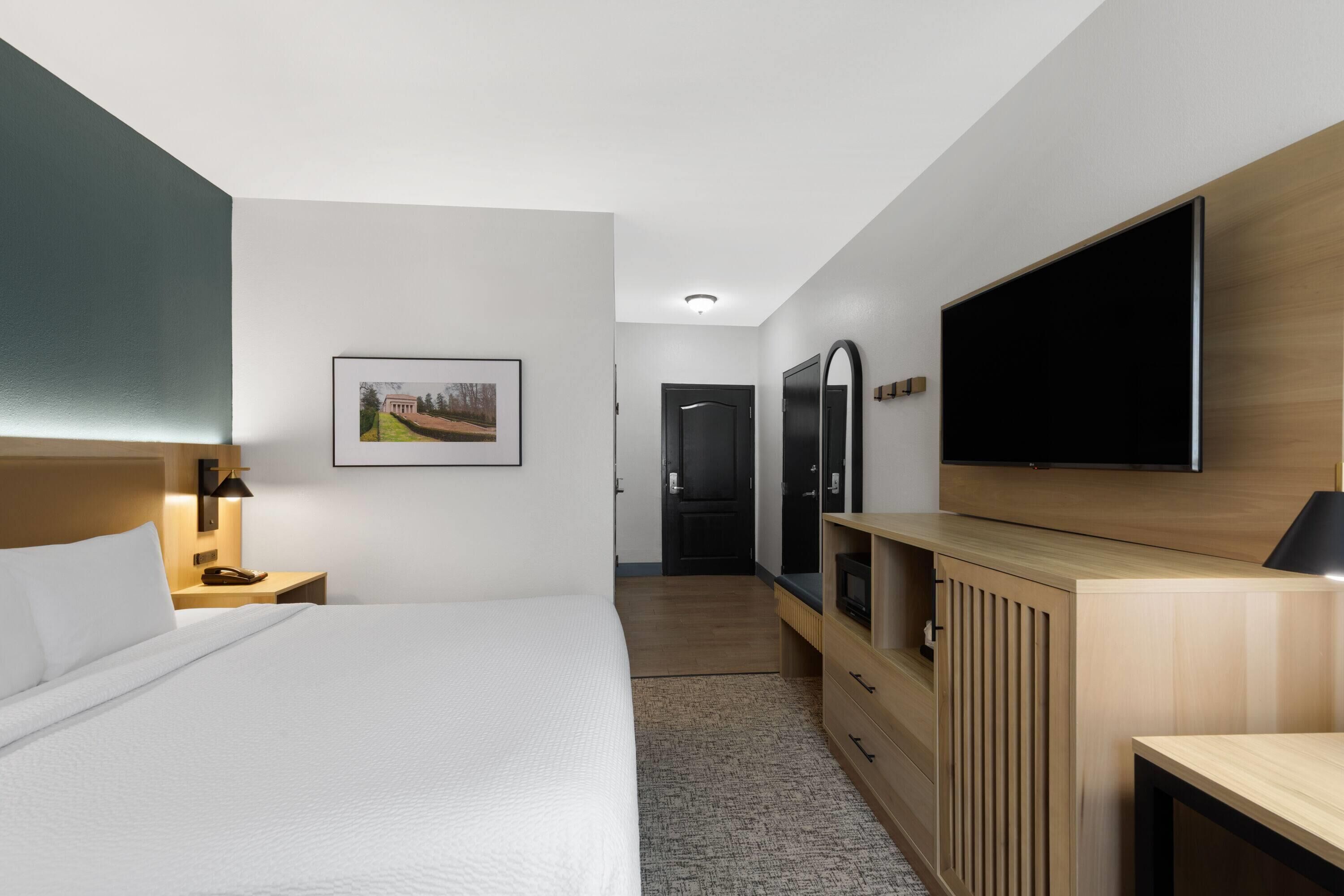 Una habitación de hotel moderna con una cama grande, un televisor de pantalla plana montado en la pared, un aparador de madera y un cuadro enmarcado.