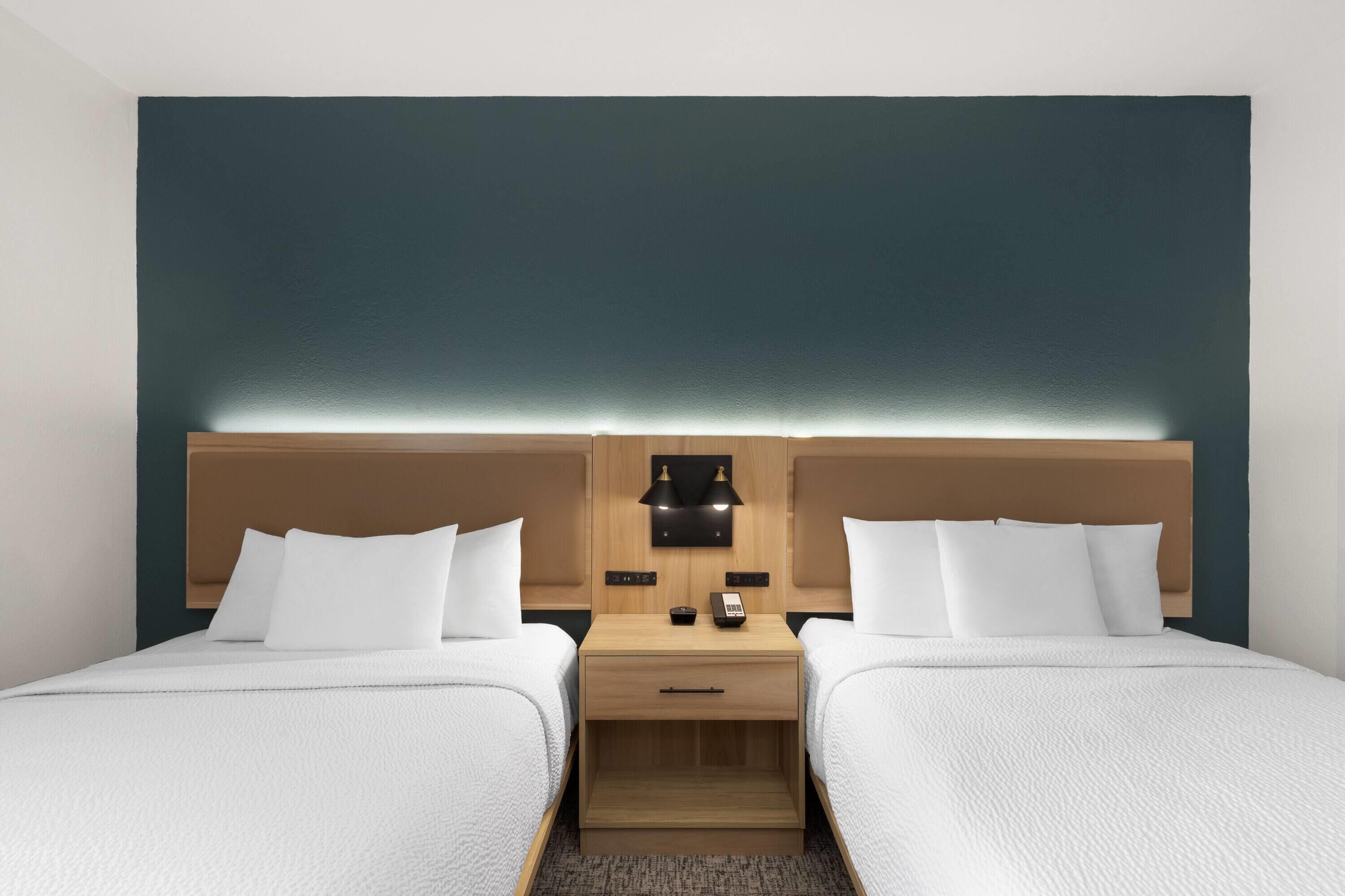 A hotel room with two beds, a wooden nightstand, a wall-mounted lamp, and a phone.