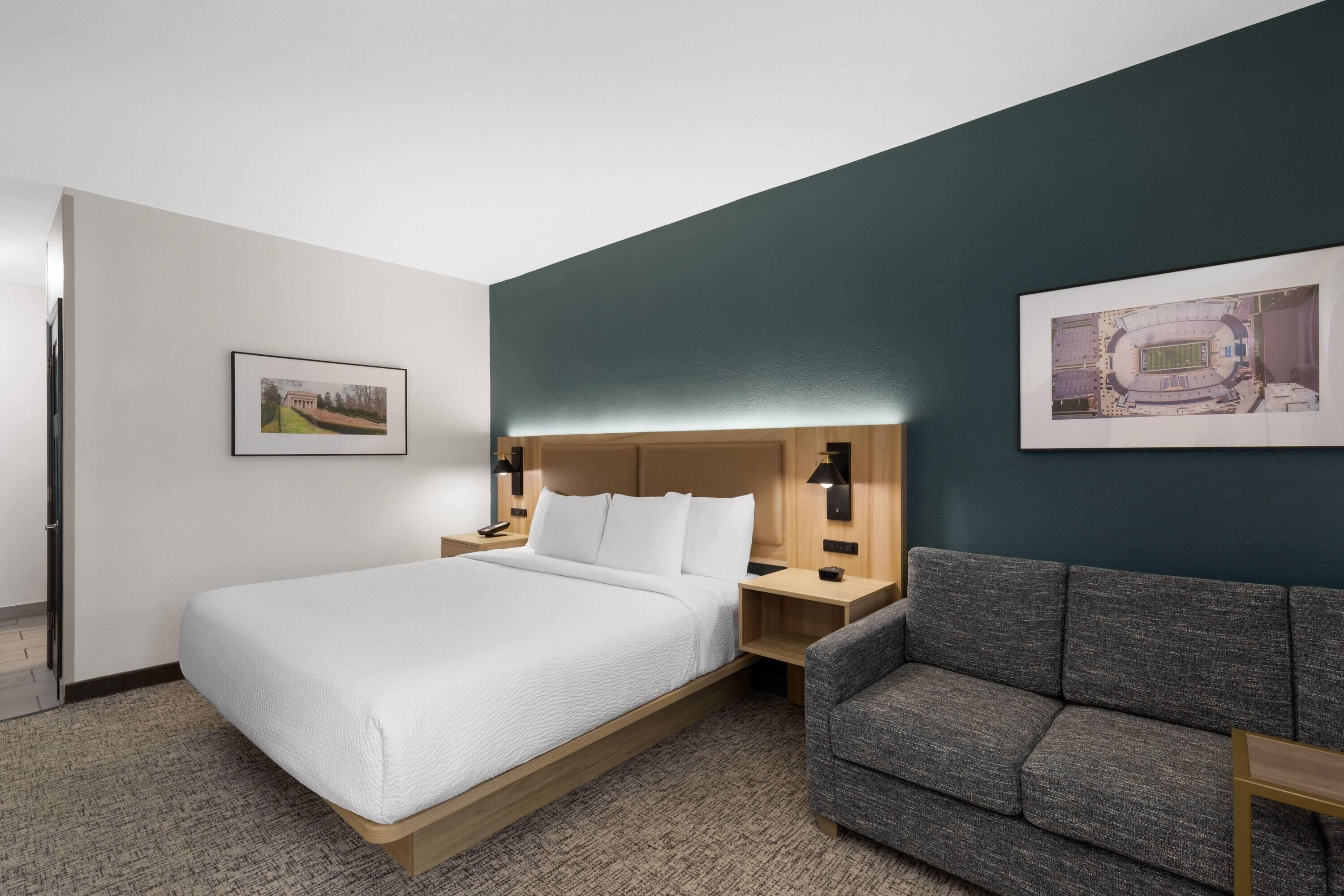 Quarto de hotel com uma cama, um sofá cinza, uma mesa de cabeceira e duas luminárias fixadas na parede.