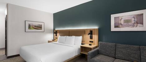 Quarto de hotel com uma cama, um sofá cinza, uma mesa de cabeceira e duas luminárias fixadas na parede.