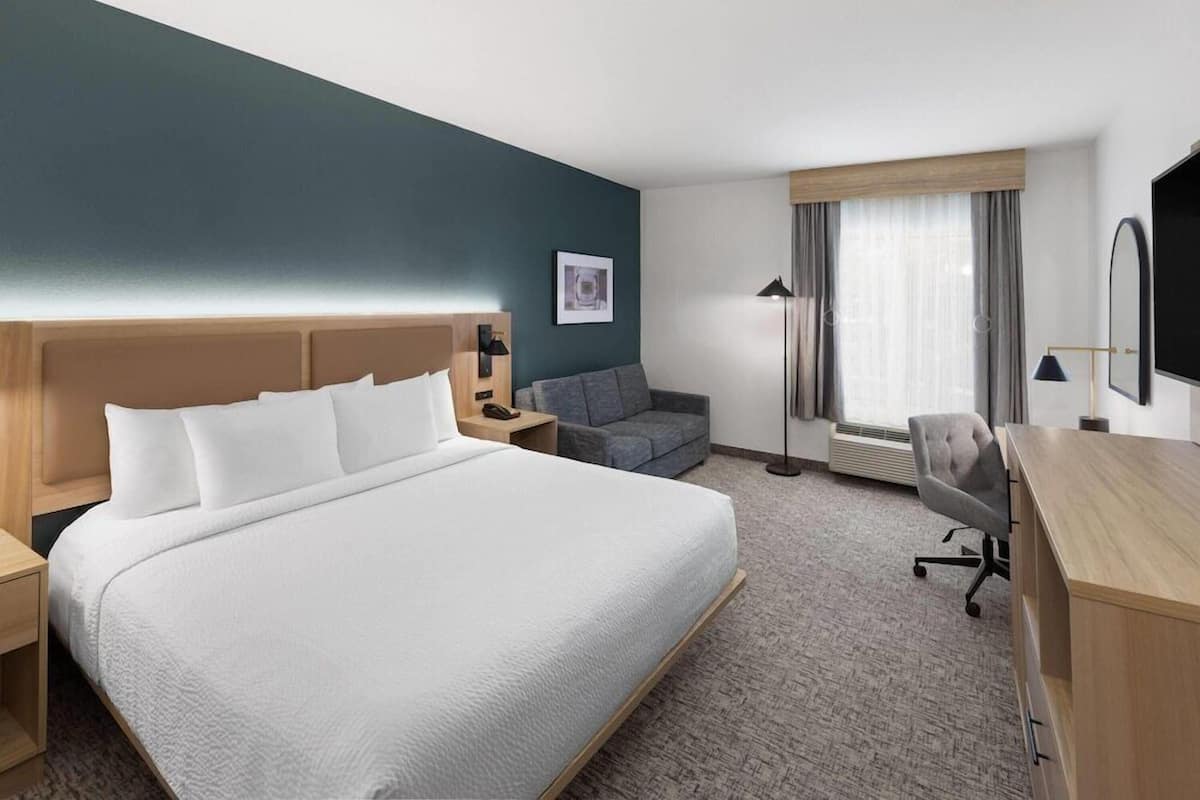Une chambre d’hôtel avec un grand lit, un bureau, une chaise, un canapé, une table de chevet, une lampe, un tableau au mur et une fenêtre avec des rideaux.