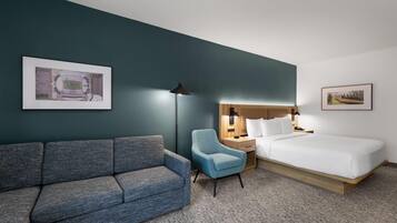 Una habitación de hotel con un sofá gris, un sillón azul, una cama, una mesita de noche y una lámpara.