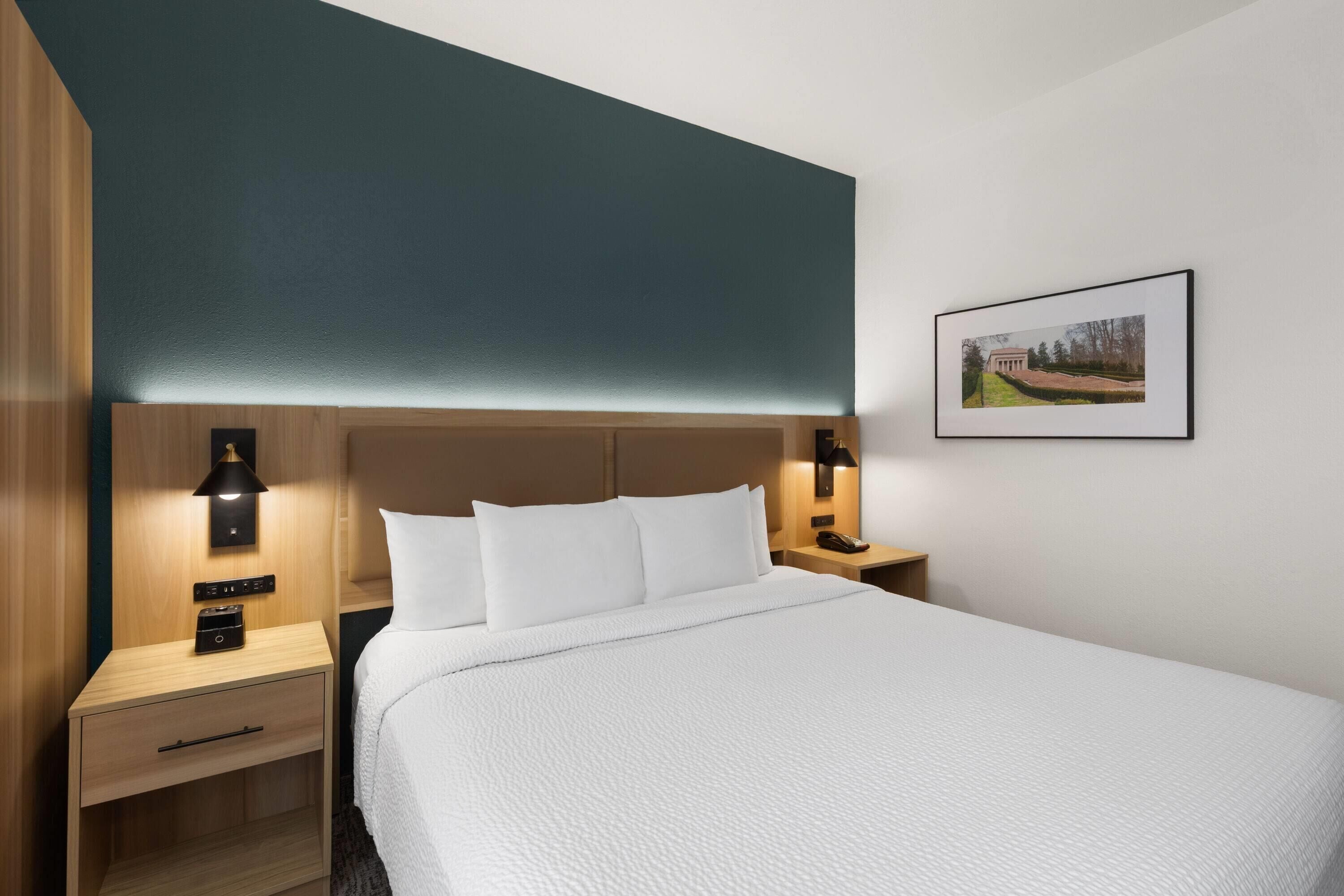 Ein Hotelzimmer mit einem großen Bett, einem Nachttisch aus Holz mit einer Lampe, einem kleinen Tischchen mit einem Telefon und einem gerahmten Bild an der Wand.