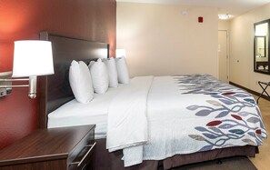 A hotel room with a bed, a nightstand, a lamp, a mirror, and a door. - Red Roof Inn Gaffney (Gaffney)