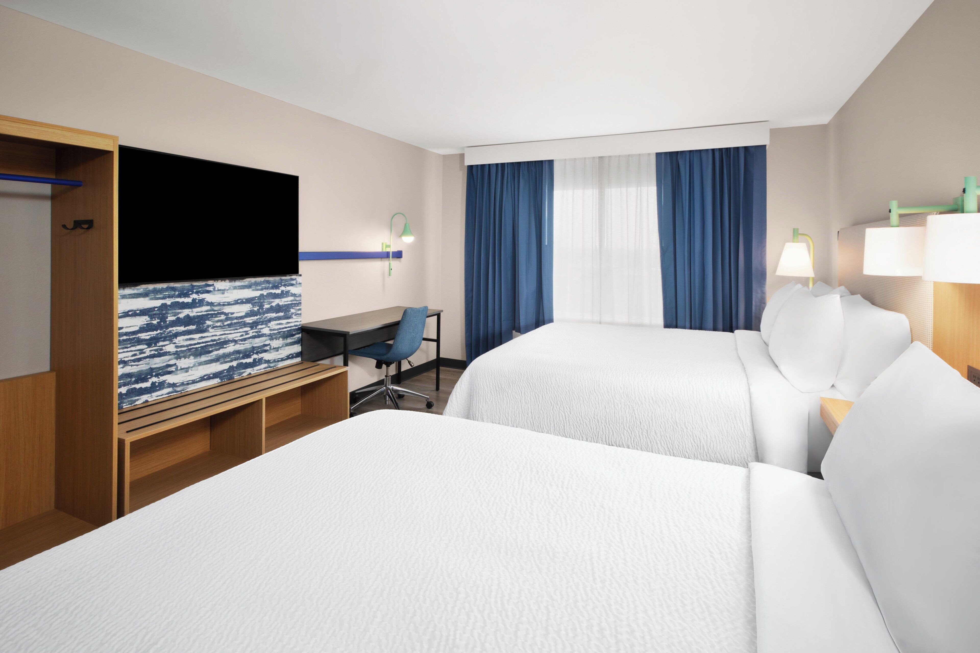 Ett hotellrum med två sängar, ett skrivbord, en stol, en TV och ett fönster med gardiner.