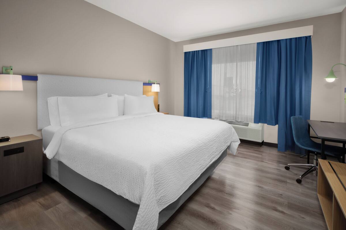 Una habitación de hotel con una cama, un escritorio, una silla, una mesita de noche, una lámpara, una ventana con cortinas y un estante en la pared.