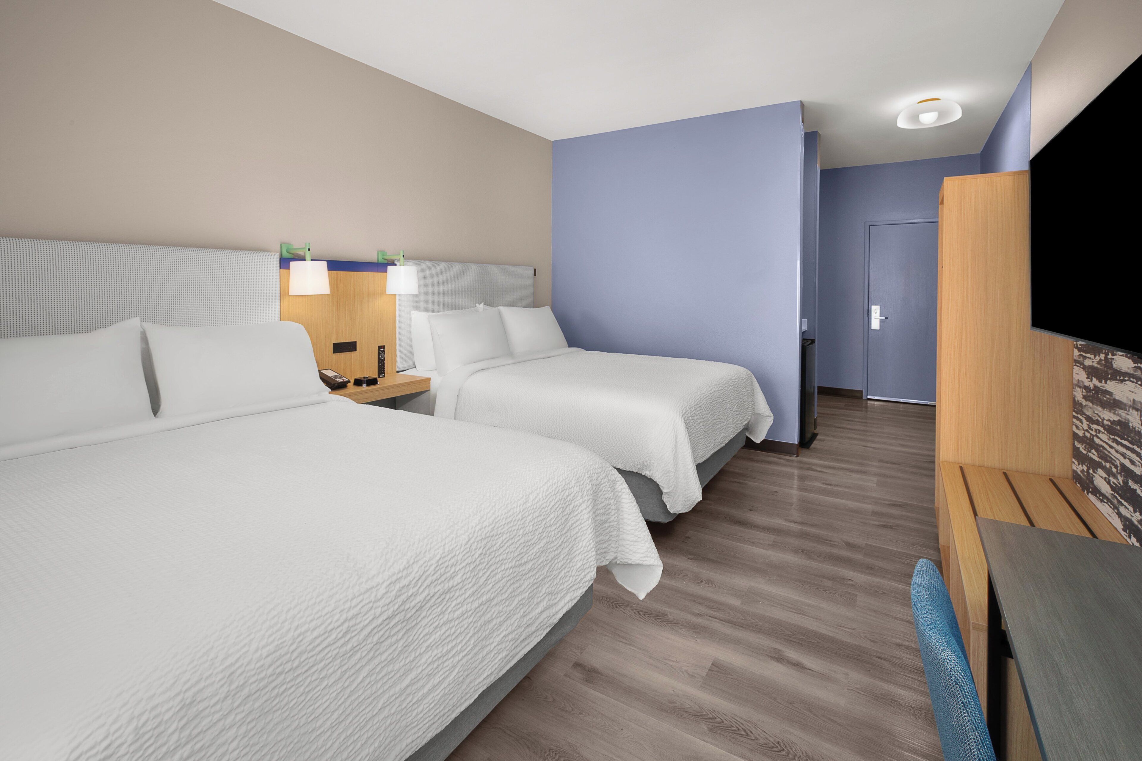 Une chambre d’hôtel avec deux lits, un bureau, une chaise, une télévision et une porte donnant sur une autre pièce.