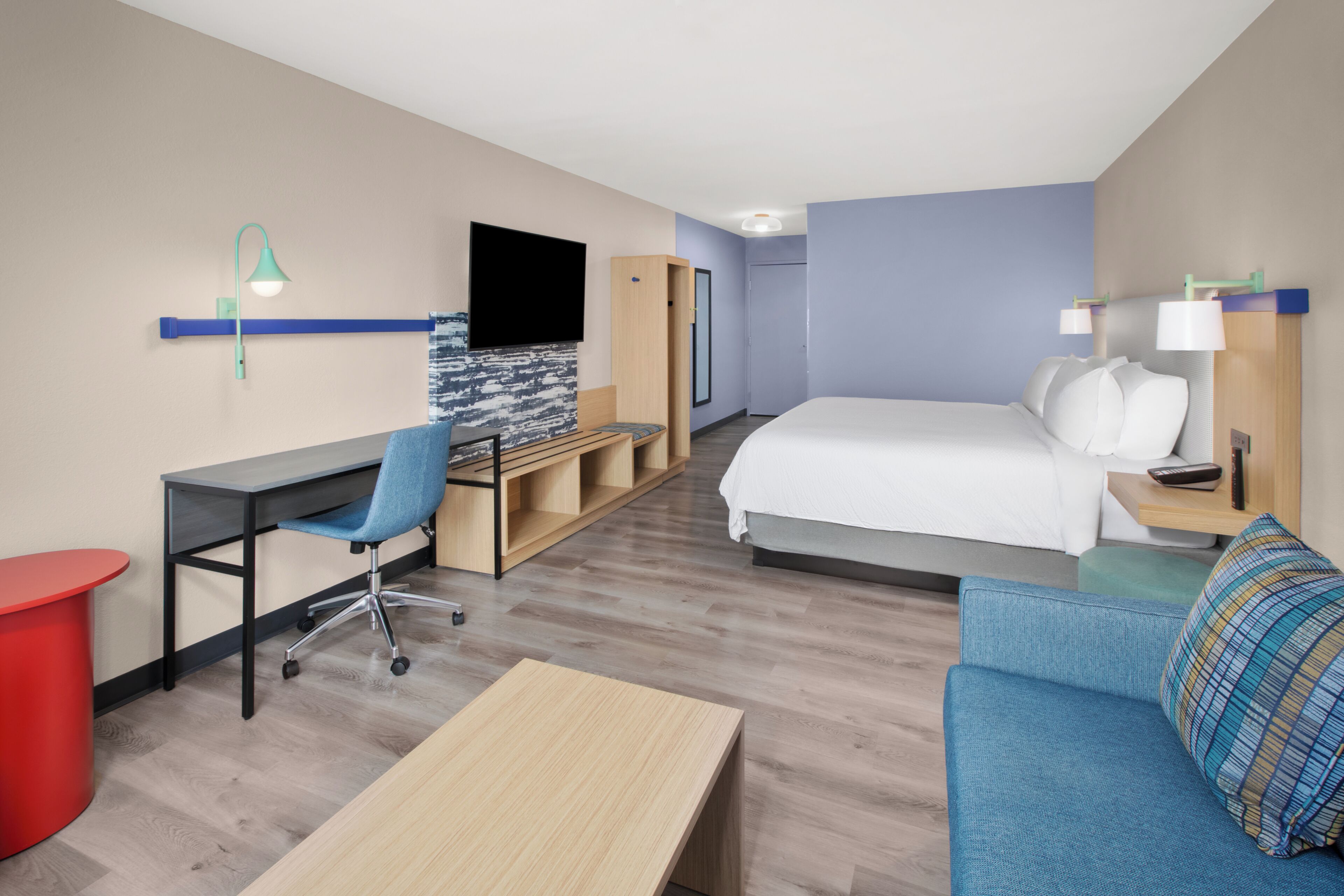 A hotel room with a bed, a desk, a chair, a TV, and a red trash bin.