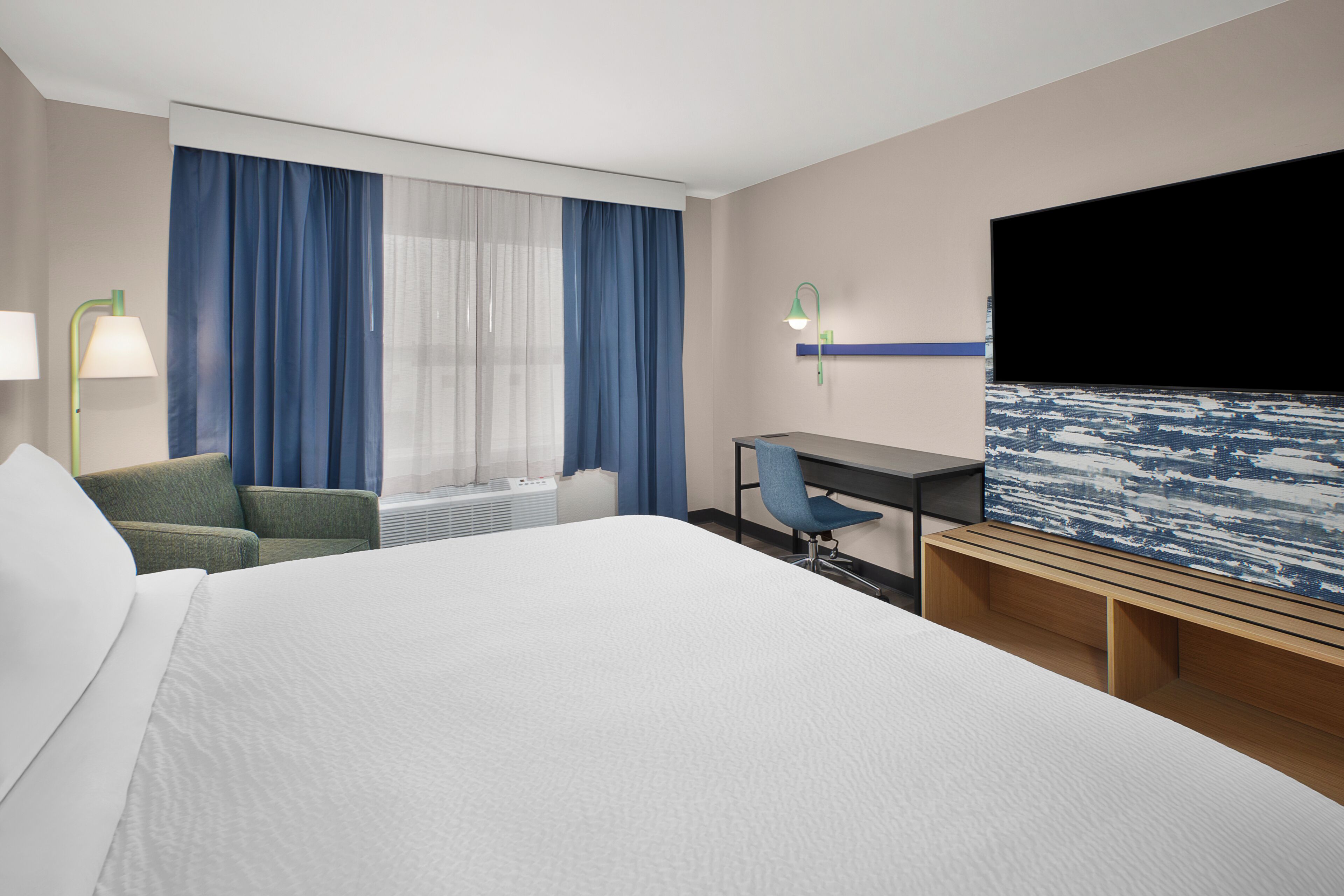 A hotel room with a large bed, a desk, a chair, a TV, and a window with curtains.