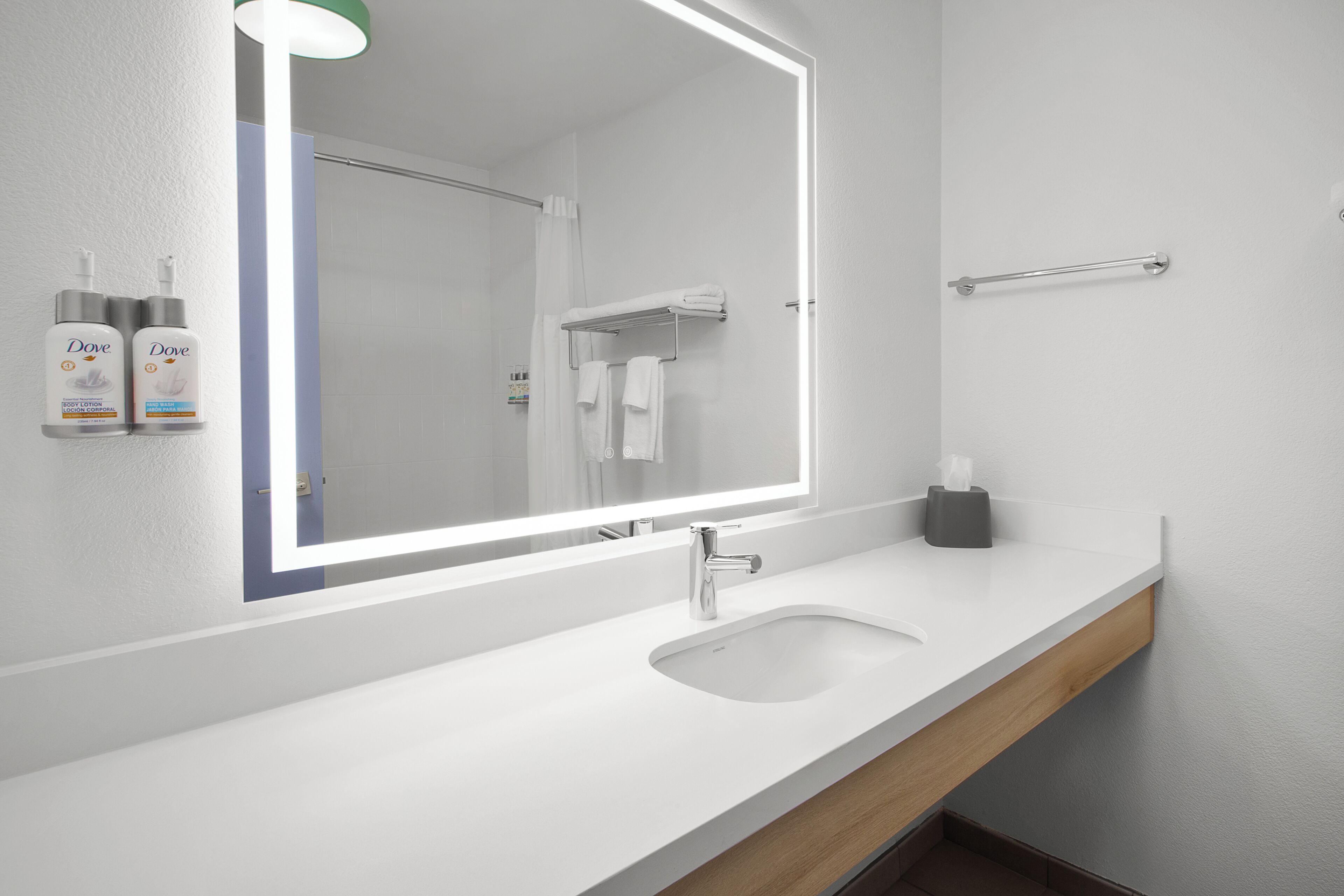 A modern bathroom with a white sink, a mirror with integrated lighting, a towel rack, and Dove lotion dispensers.