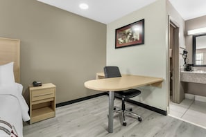A hotel room with a bed, a desk with a chair, a nightstand, a framed picture on the wall, and a bathroom visible through an open door.