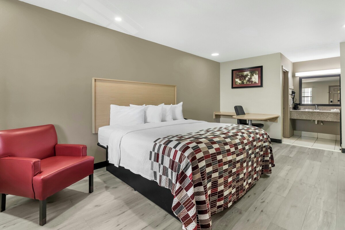 A hotel room with a bed, a red chair, a desk, and a bathroom with a mirror.