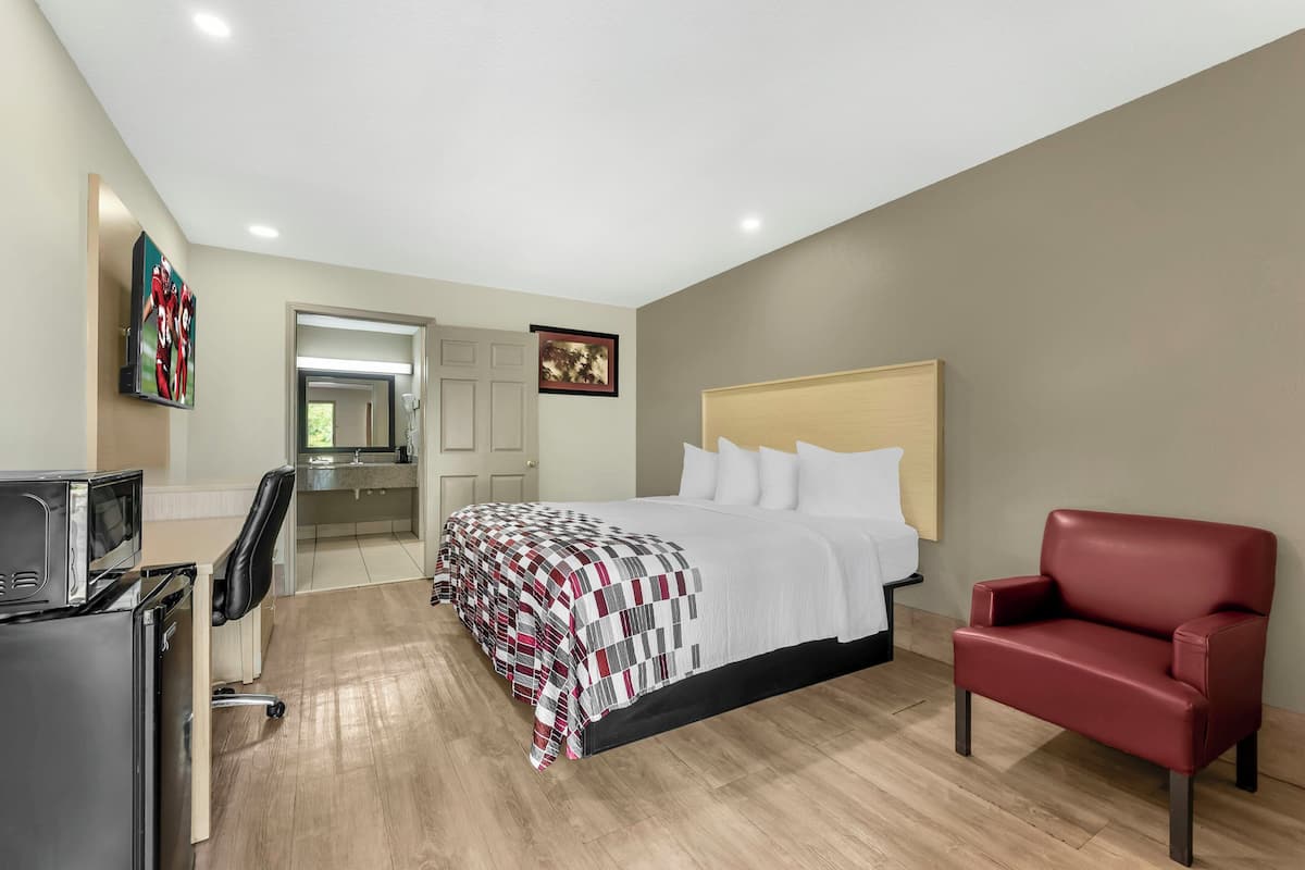 Une chambre d’hôtel avec un lit, une chaise rouge, un bureau avec un ordinateur et une télévision.