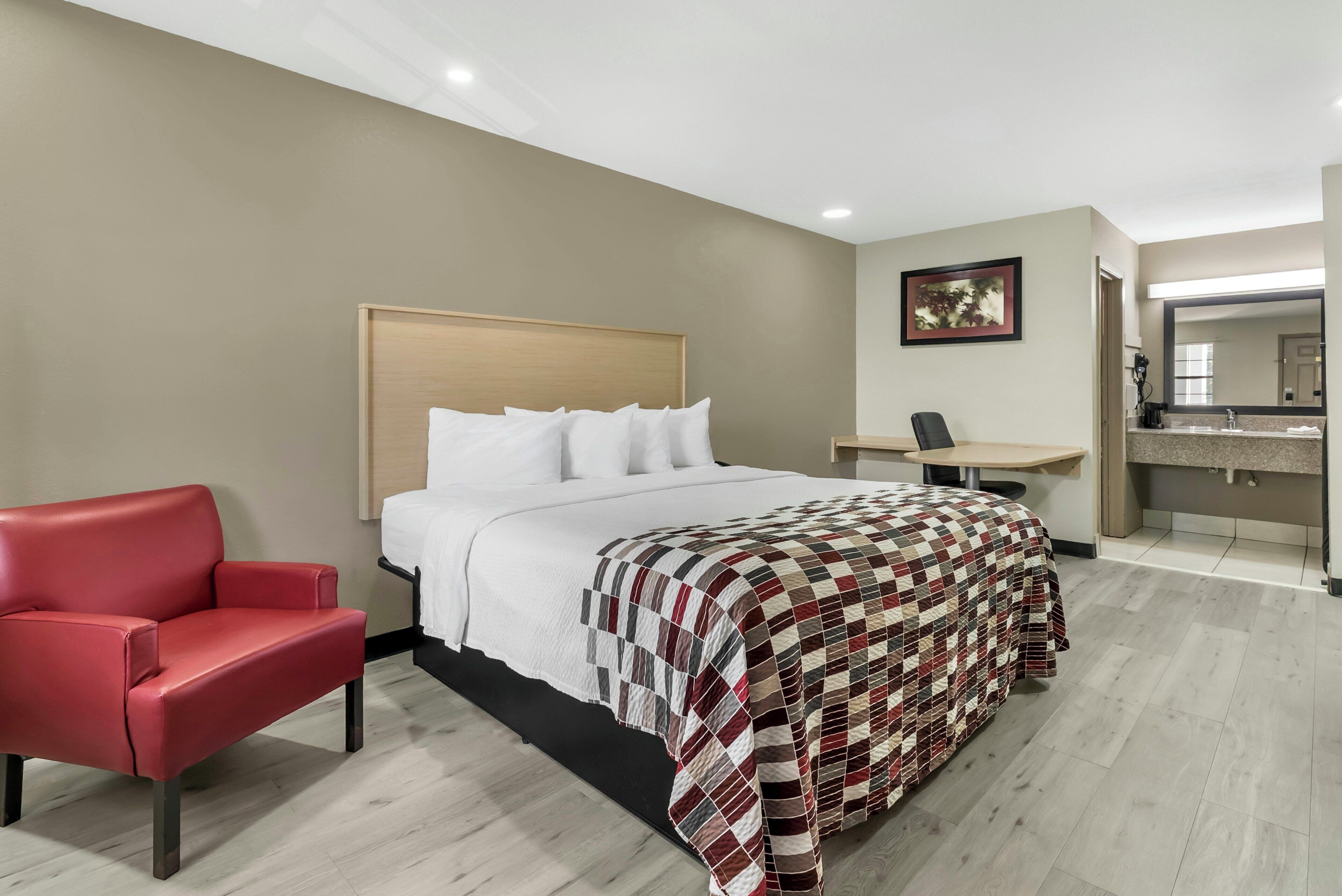 A hotel room with a bed, a red chair, a desk, and a bathroom with a mirror.