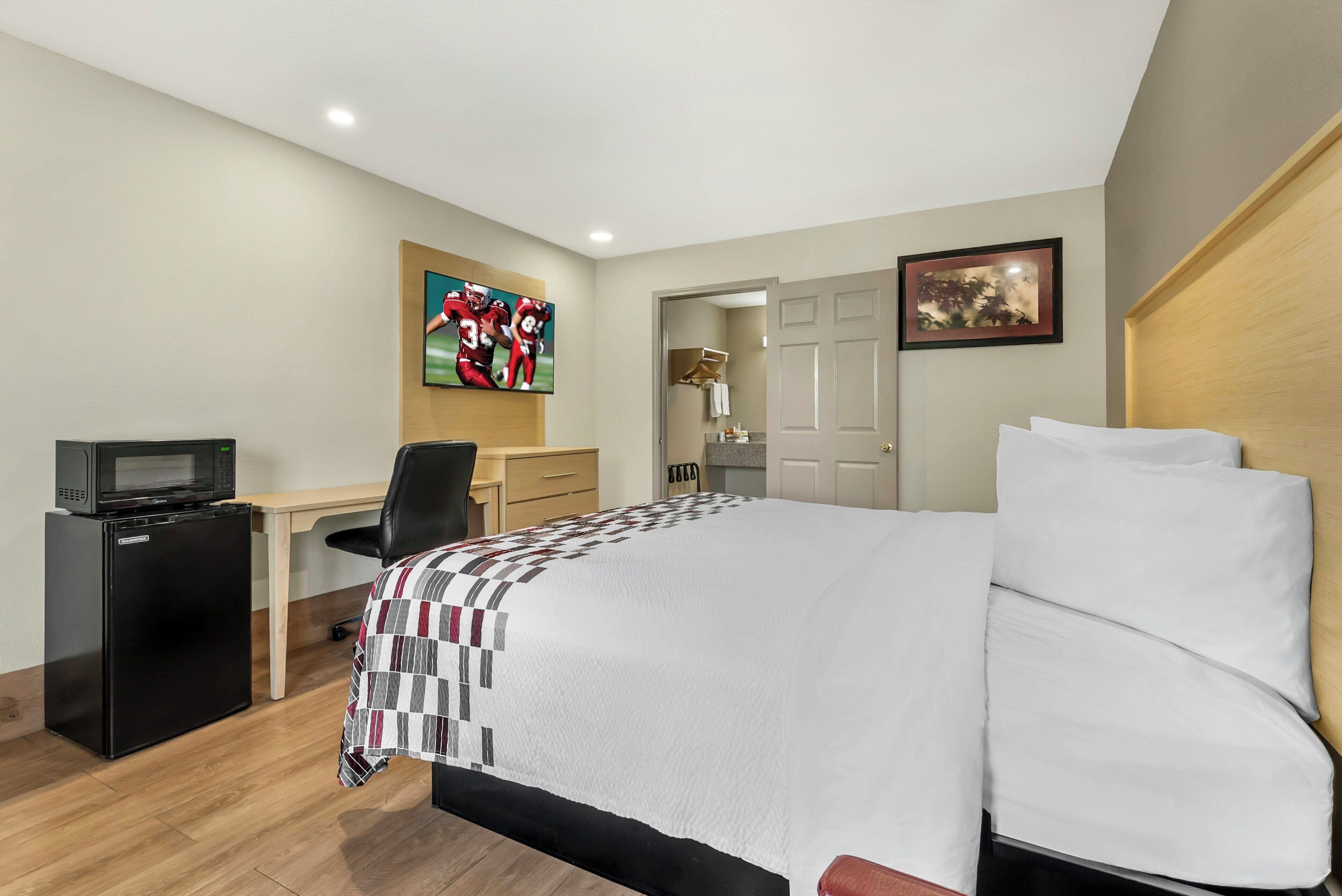 A hotel room with a bed, a desk with a computer, a refrigerator, a TV, and a door leading to a bathroom.