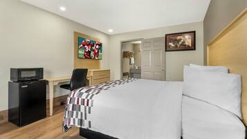 A hotel room with a bed, a desk with a computer, a refrigerator, a TV, and a door leading to a bathroom.
